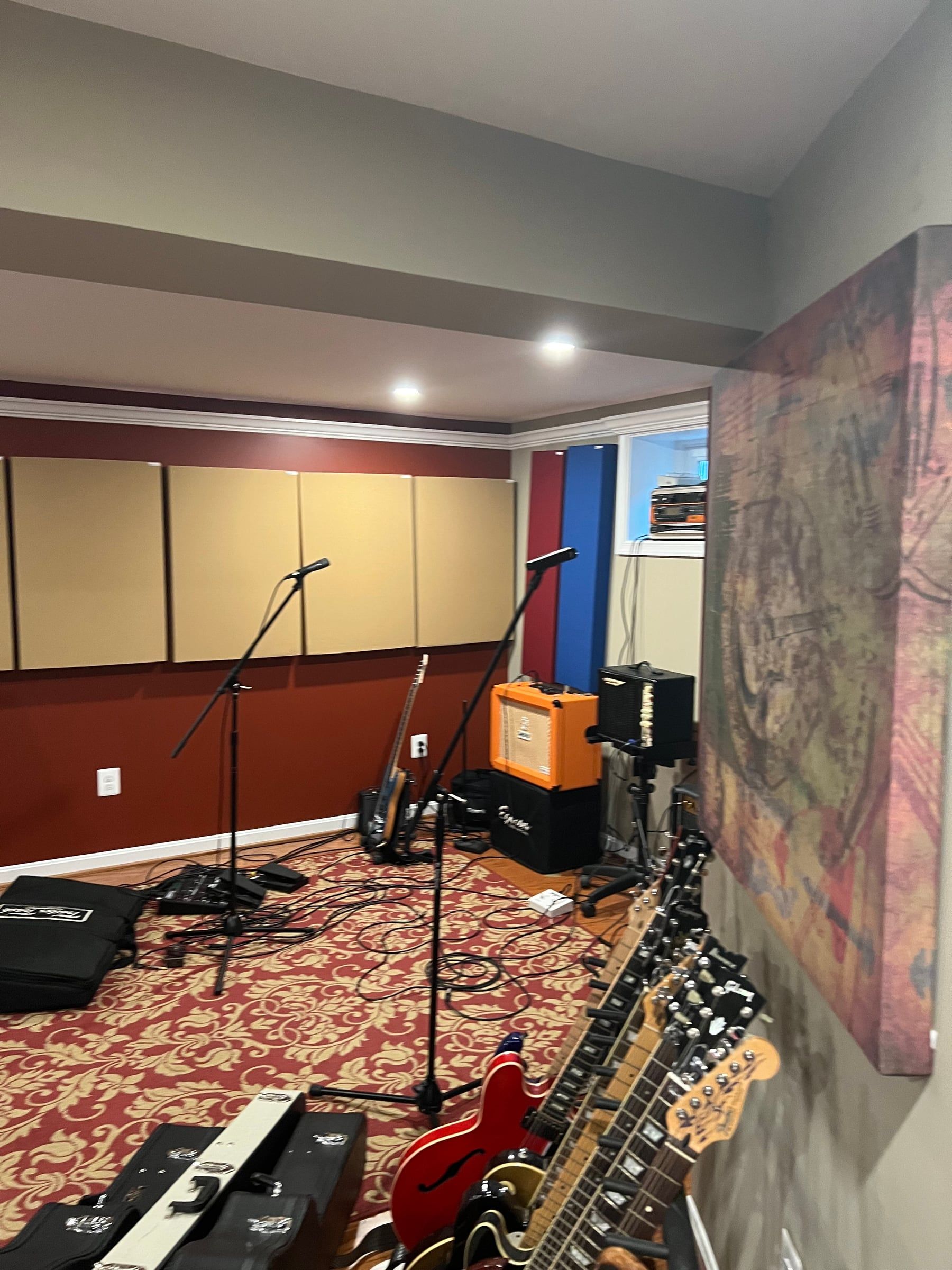 The live room: microphone stands, an orange guitar amp and a combo amp, cables across a patterned rug, and a row of electric guitars on stands